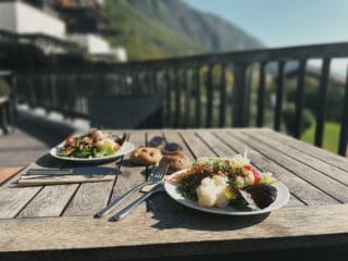 Veganer Teller mit frischem Gem&uuml;se und Salat auf einem Holztablett, mit Bergpanorama im Hintergrund.