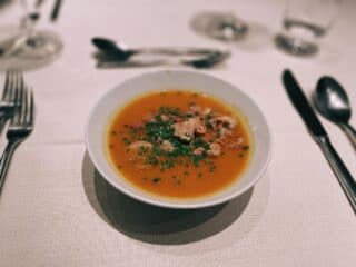 Vegane Suppe in einer wei&szlig;en Sch&uuml;ssel mit Kr&auml;utern, serviert auf einem Esstisch.