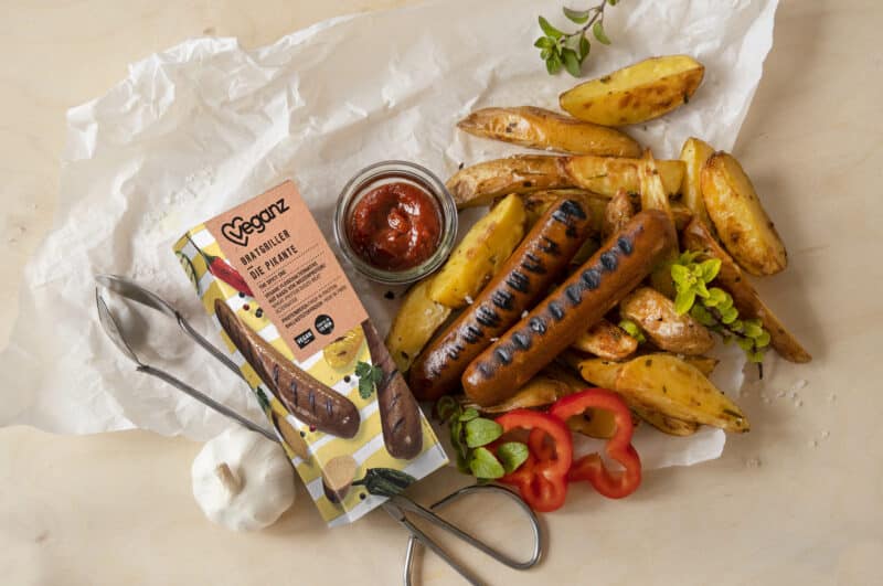 Auf einem weißen Backpapier liegen fertig gegrillte Wurste von veganz. Daneben die Verpackung der Würste . Neben den Würsten liegen gebackene Kartoffelecken und Paprika