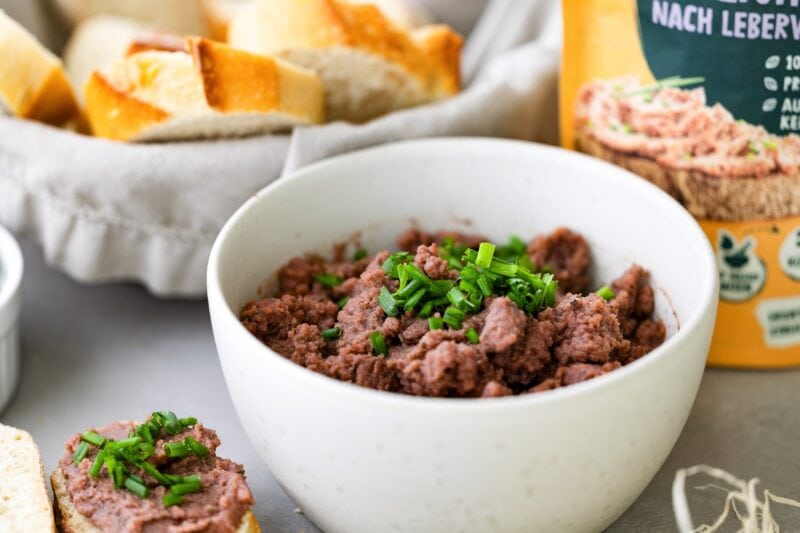 Schale mit veganer Leberwurst darin, die mit Schnittlauch dekoriert ist. Im Hintergrund geschnittenes Baguette und die Verpackung der Leberwurst
