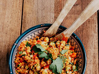 Runde Schale mit Couscoussalat der mit Gurke, Tomaten und Paprika gemacht wurde. In der Schale befindet sich Salatbesteck aus Holz.