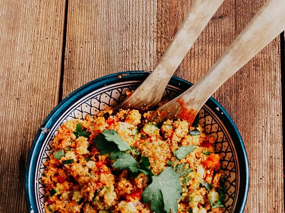 Runde Schale mit Couscoussalat der mit Gurke, Tomaten und Paprika gemacht wurde. In der Schale befindet sich Salatbesteck aus Holz.