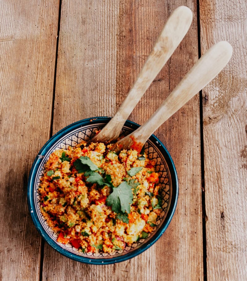Runde Schale mit Couscoussalat der mit Gurke, Tomaten und Paprika gemacht wurde. In der Schale befindet sich Salatbesteck aus Holz.
