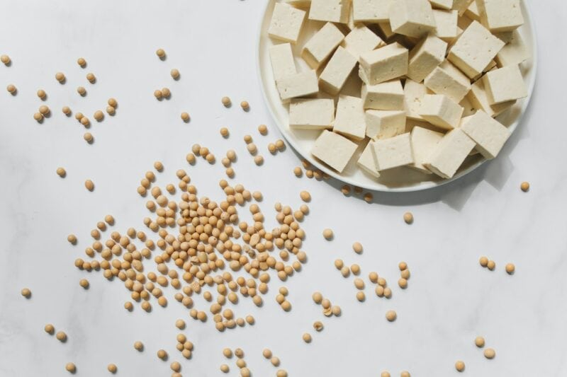 weiße Oberfläche mit einem runden Teller auf dem gewürfeltes Tofu liegt, daneben Soja Bohnen verstreut