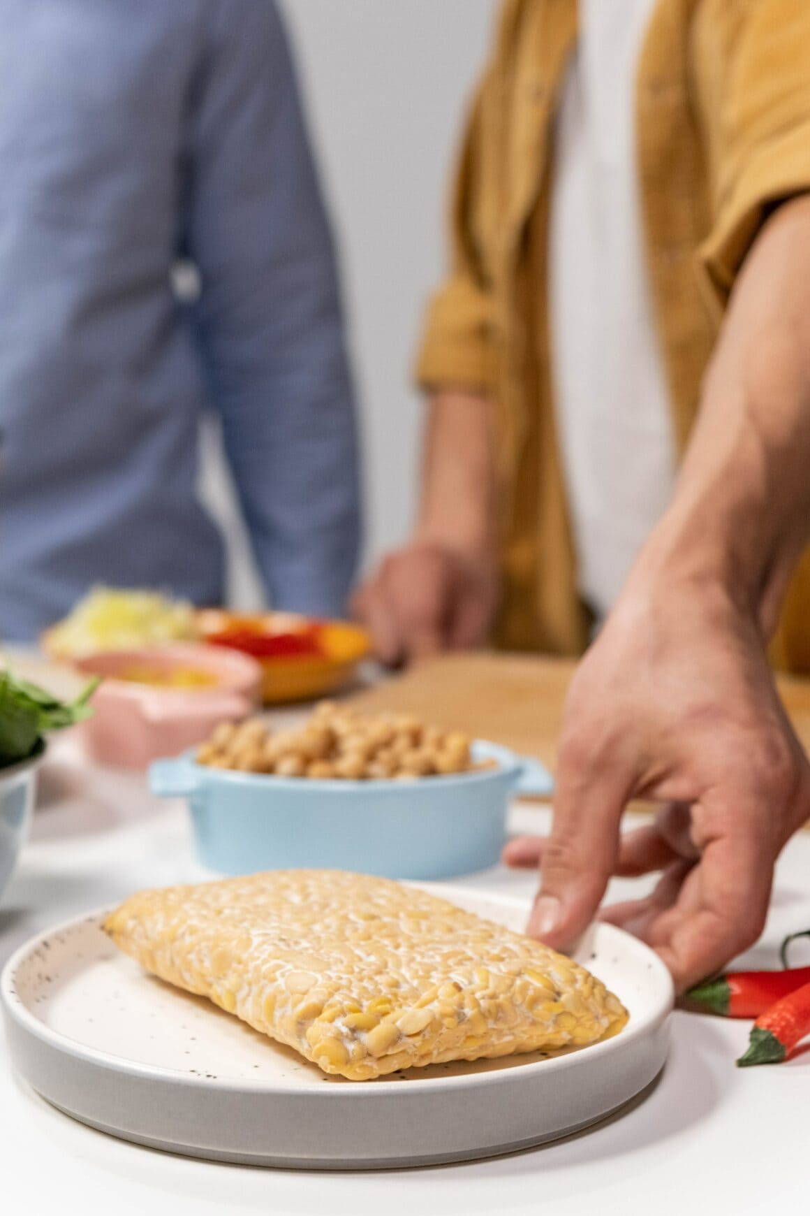 runder weißer Teller auf dem Tempeh liegt, im Hintergrund sind weitere runde Schalen mit Lebensmitteln zu sehen