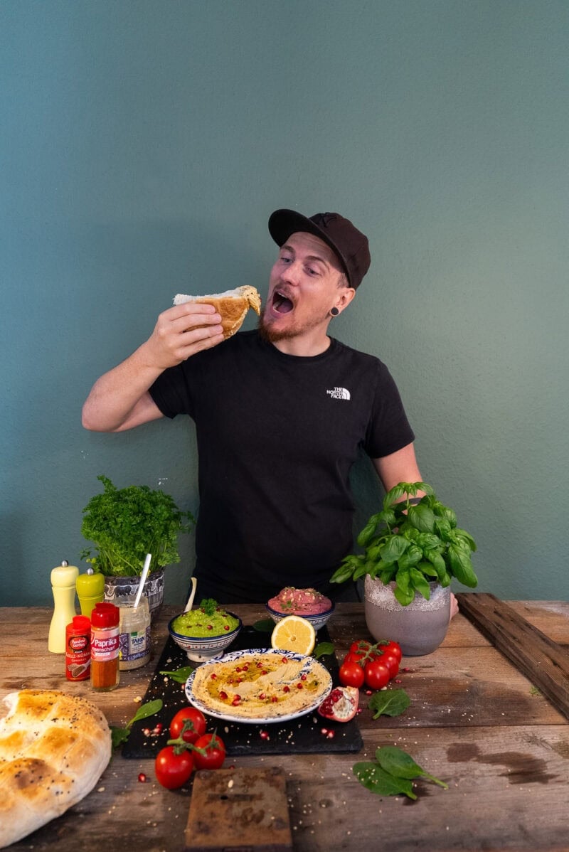 Yannick steht mit offenem Mund vor einem Tisch und Hält ein Brot mit Hummus in der Hand. Vor Ihm stehen 3 Schalen mit Hummus und ein Fladenbrot