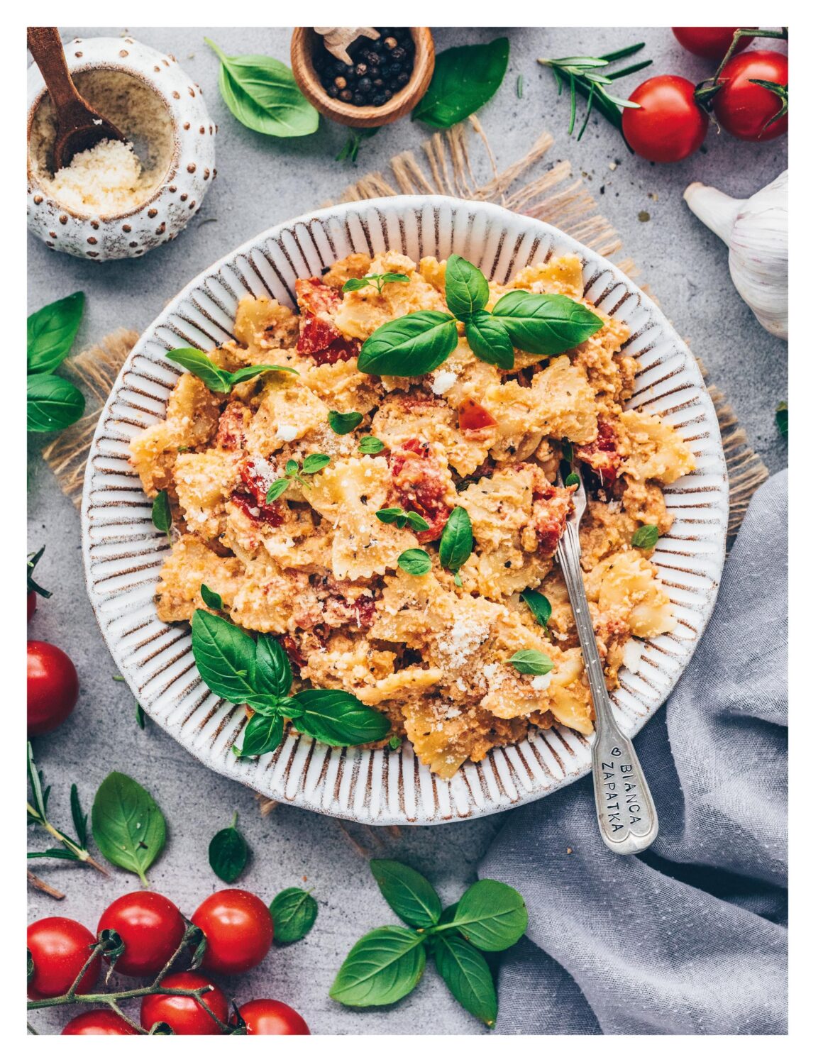 Vegane Baked Feta Pasta mit Kirschtomaten und frischem Basilikum auf einem Teller, umgeben von Tomaten und Kräutern