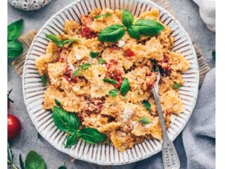 Vegane Baked Feta Pasta mit Kirschtomaten und frischem Basilikum auf einem Teller, umgeben von Tomaten und Kräutern