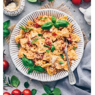 Vegane Baked Feta Pasta mit Kirschtomaten und frischem Basilikum auf einem Teller, umgeben von Tomaten und Kräutern