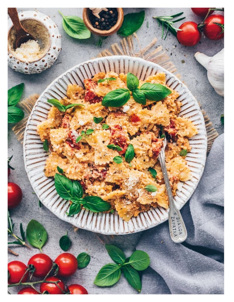 Vegane Baked Feta Pasta mit Kirschtomaten und frischem Basilikum auf einem Teller, umgeben von Tomaten und Kräutern