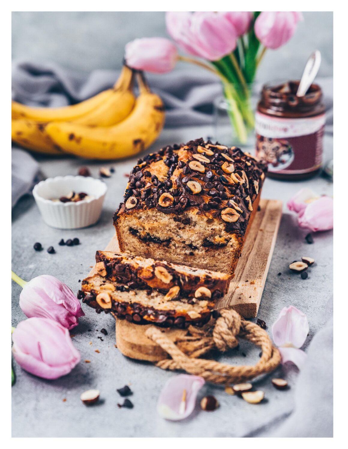 Veganes Schoko-Bananenbrot mit Haselnüssen und Schokostückchen auf Holzbrett, umgeben von Bananen, Tulpen und Schokoaufstrich