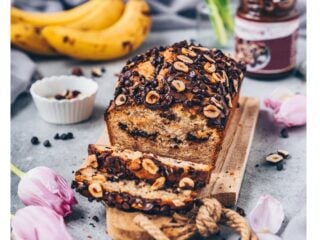 Veganes Schoko-Bananenbrot mit Haselnüssen und Schokostückchen auf Holzbrett, umgeben von Bananen, Tulpen und Schokoaufstrich