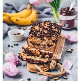 Veganes Schoko-Bananenbrot mit Haselnüssen und Schokostückchen auf Holzbrett, umgeben von Bananen, Tulpen und Schokoaufstrich