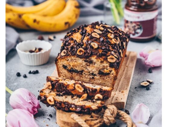 Veganes Schoko-Bananenbrot mit Haselnüssen und Schokostückchen auf Holzbrett, umgeben von Bananen, Tulpen und Schokoaufstrich