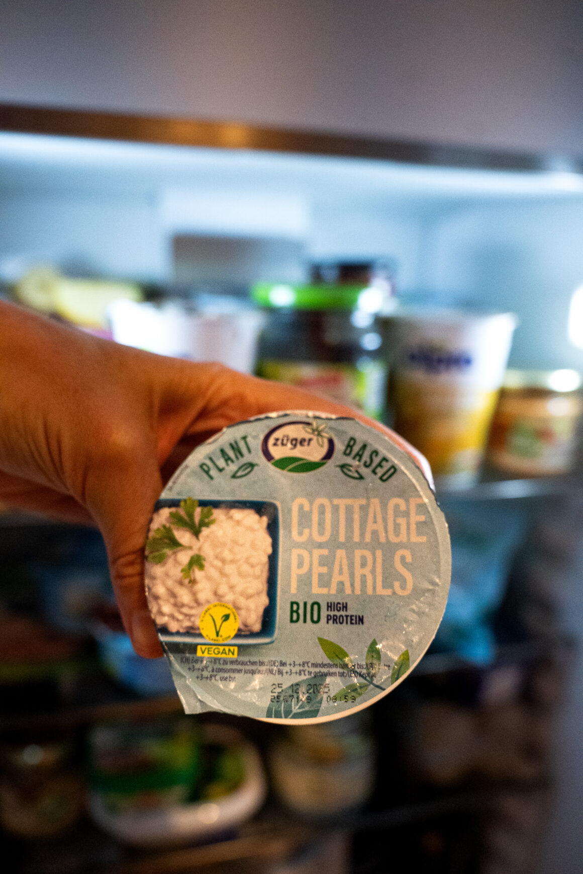 Hand hält Züger Plant-Based Cottage Pearls Bio High Protein Becher vor geöffnetem Kühlschrank mit verschiedenen Lebensmitteln im Hintergrund