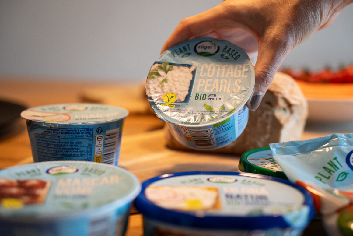 Hand hält Züger Plant-Based Cottage Pearls Bio High Protein Becher, im Hintergrund weitere vegane Produkte der Züger Linie wie MascarVone und Filona auf Holztisch