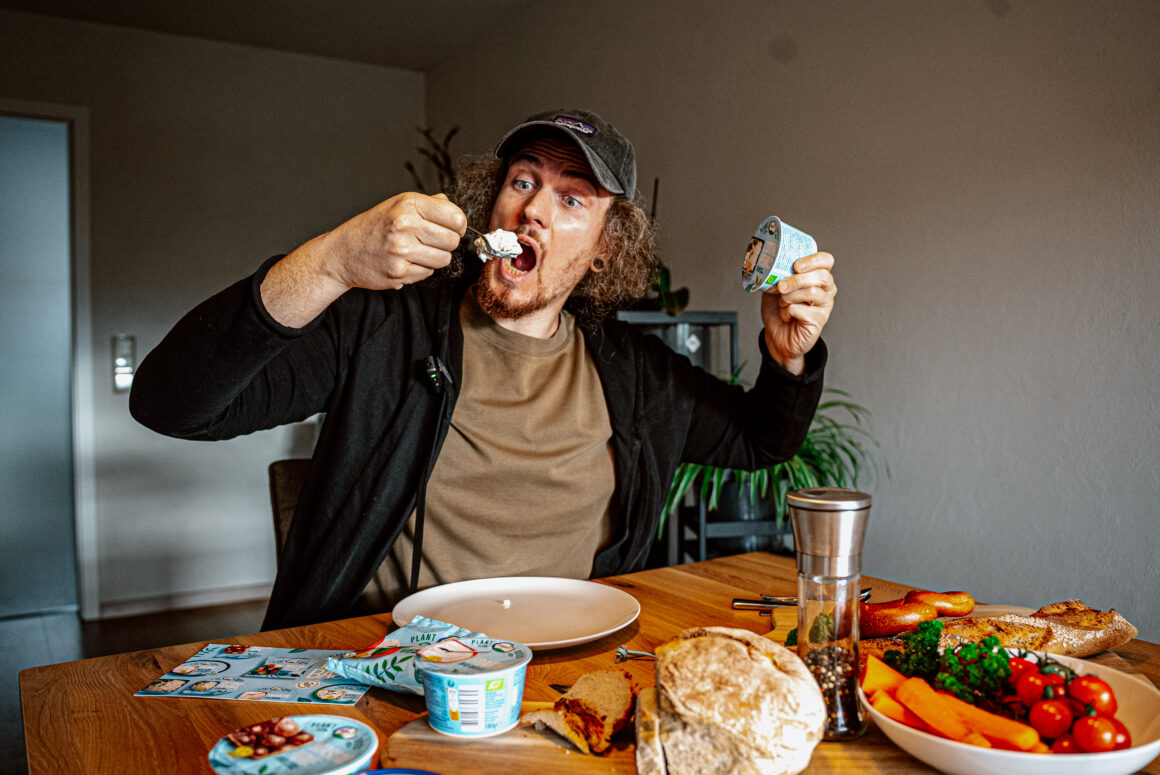 Mann mit erstauntem Gesichtsausdruck hält Züger Plant-Based MozzaVella und Cottage Pearls hoch, vor ihm auf dem Holztisch das komplette vegane Sortiment mit frischem Gemüse und Brot