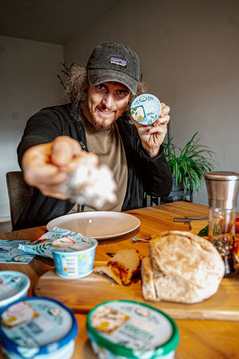 Veganer Käse in der Hand, umgeben von verschiedenen veganen Käseprodukten auf dem Tisch.