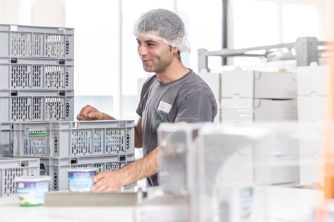 Lächelnder Züger Mitarbeiter mit Haarnetz beim Verpacken von Plant-Based Produkten in Transportkisten in der modernen Produktionshalle in Oberbüren