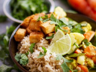 Veganes Frühlingscurry mit knusprigem Erdnusstofu, Naturreis, frischem Koriander und Limette in einer dunklen Bowl
