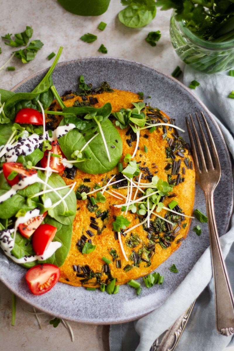 Veganes Kichererbsen-Omelette mit frischen Kräutern, Babyspinat, Cherrytomaten, Sprossen und Sojajoghurt auf grauem Keramikteller
