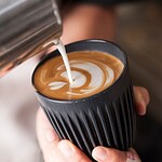 Ist Kaffee vegan? Die ehrliche Stolperfallen-Liste plus die besten Pflanzendrink-Pairings 2026 Barista pouring milk to create latte art in a black reusable coffee cup