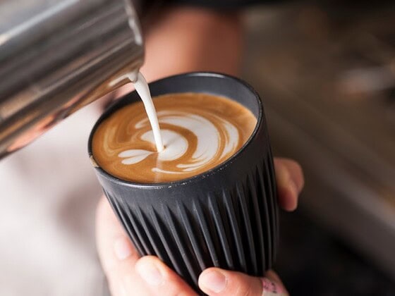Pflanzliche Milch wird aus einem Metallkännchen in einen schwarzen Becher mit Kaffee gegossen und formt Latte-Art