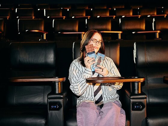 Billie Eilish im Kinosessel mit zwei Tafeln der veganen iChoc Limited Edition zum Kinostart von Hit Me Hard and Soft