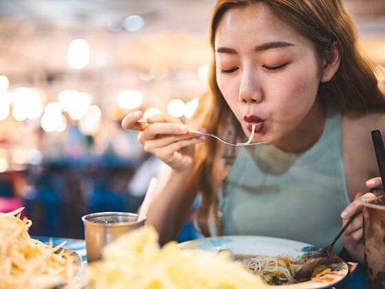 Person mit Rucksack vor exotischer Streetfood-Szene, vegan reisen in Asien mit Buddha Bowl in der Hand