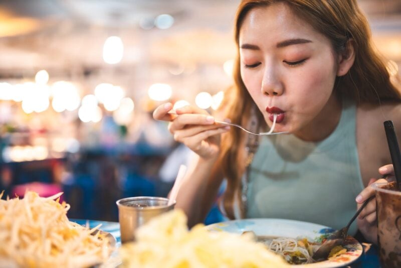 Person mit Rucksack vor exotischer Streetfood-Szene, vegan reisen in Asien mit Buddha Bowl in der Hand