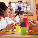 Großbritannien reformiert das Schulessen – und Hülsenfrüchte spielen die Hauptrolle Lachende Schulkinder essen gemeinsam in einer Schulkantine mit bunten Bechern und Tellern