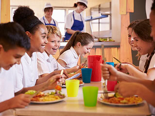 Lachende Schulkinder essen gemeinsam in einer Schulkantine mit bunten Bechern und Tellern
