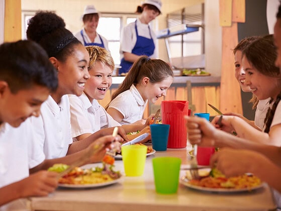 Lachende Schulkinder essen gemeinsam in einer Schulkantine mit bunten Bechern und Tellern