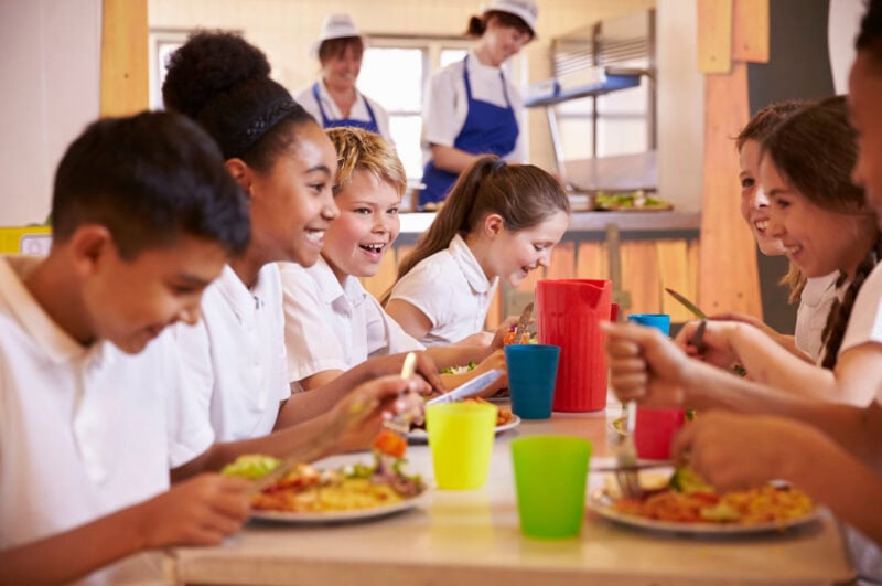 Lachende Schulkinder essen gemeinsam in einer Schulkantine mit bunten Bechern und Tellern