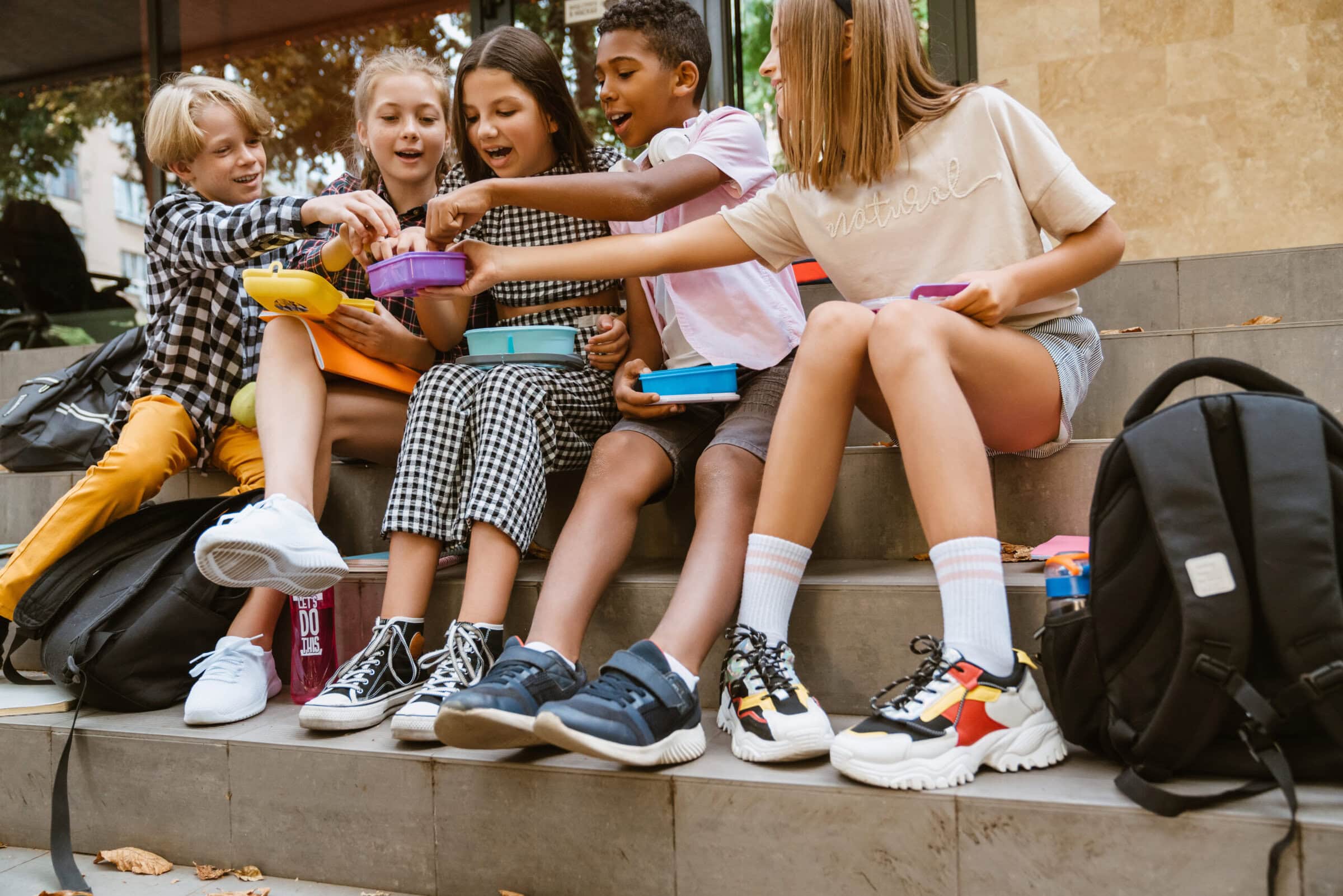 Schulkinder sitzen auf einer Treppe und essen aus bunten Lunchboxen