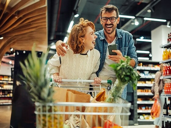Paar mit Smartphone und vollem Einkaufswagen - veganer Wocheneinkauf