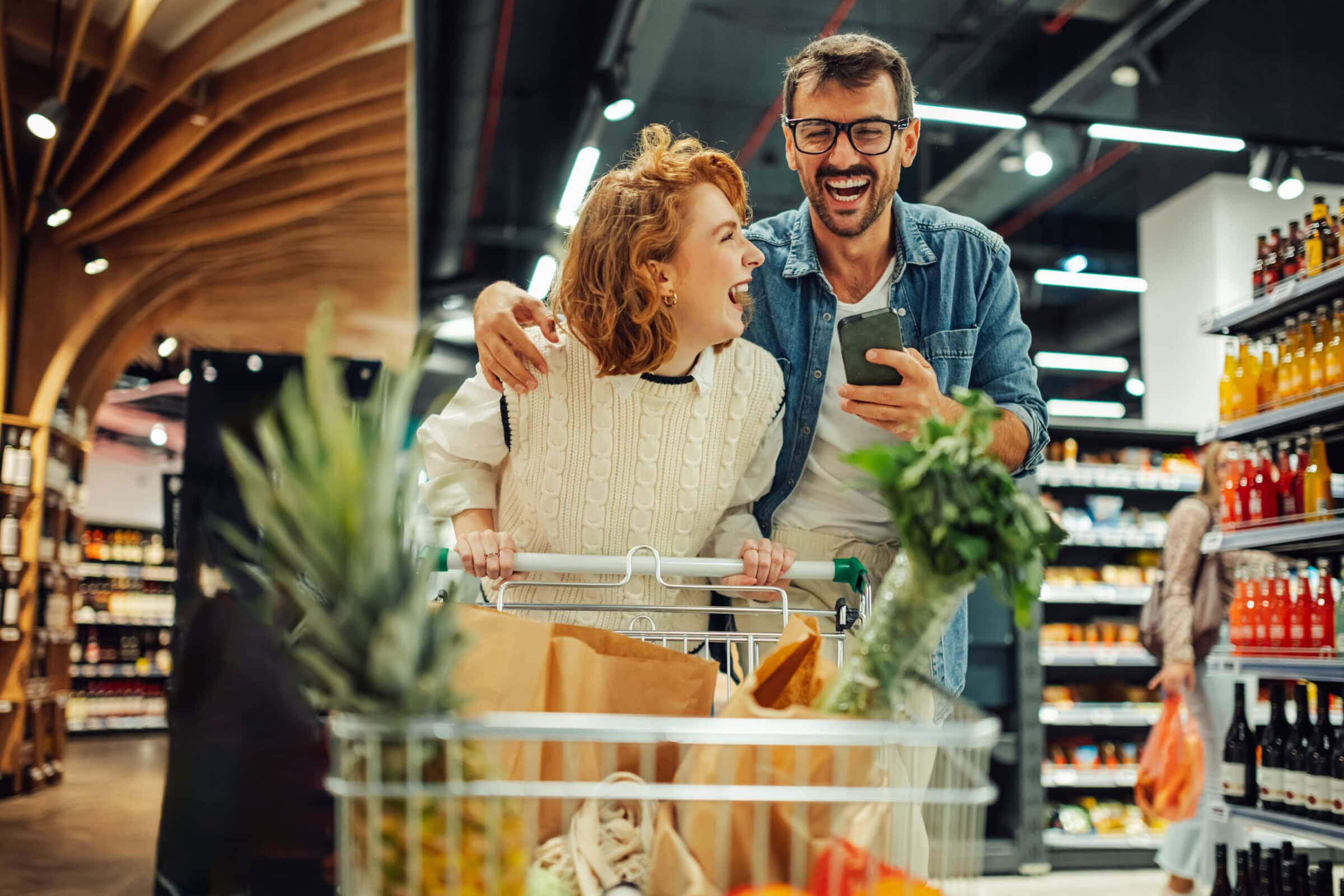 Paar mit Smartphone und vollem Einkaufswagen - veganer Wocheneinkauf