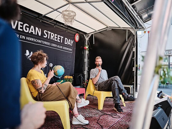 Zwei Menschen im Gespräch auf gelben Stühlen auf der Bühne des Vegan Street Day vor einem Banner
