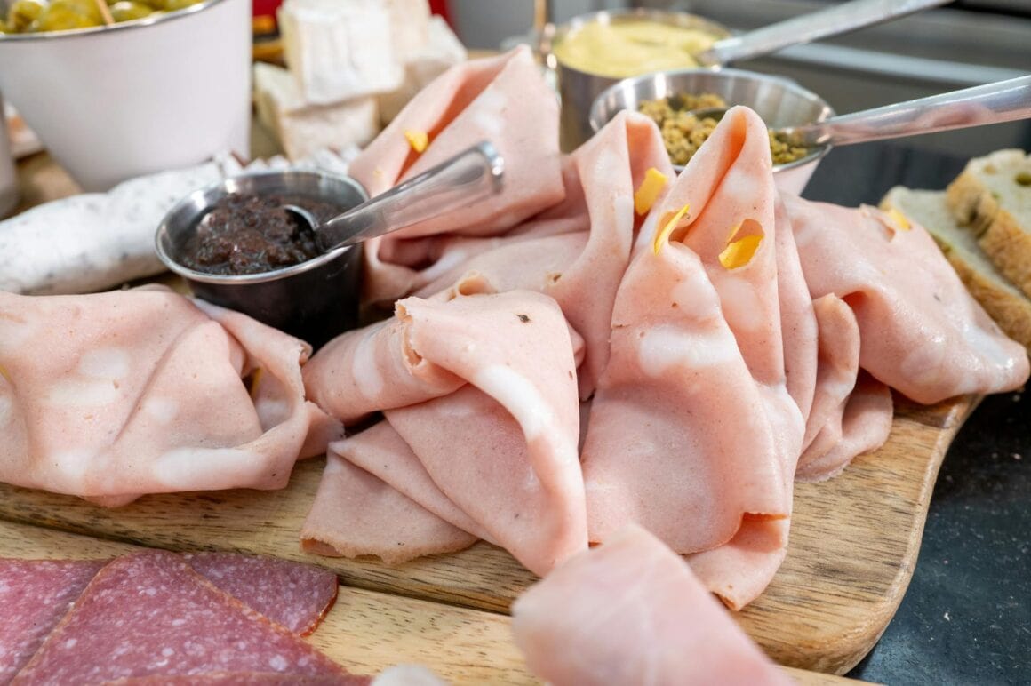 Veganer Aufschnitt in verschiedenen Sorten auf Holzbrett, daneben Brötchen, Senf und Gurke