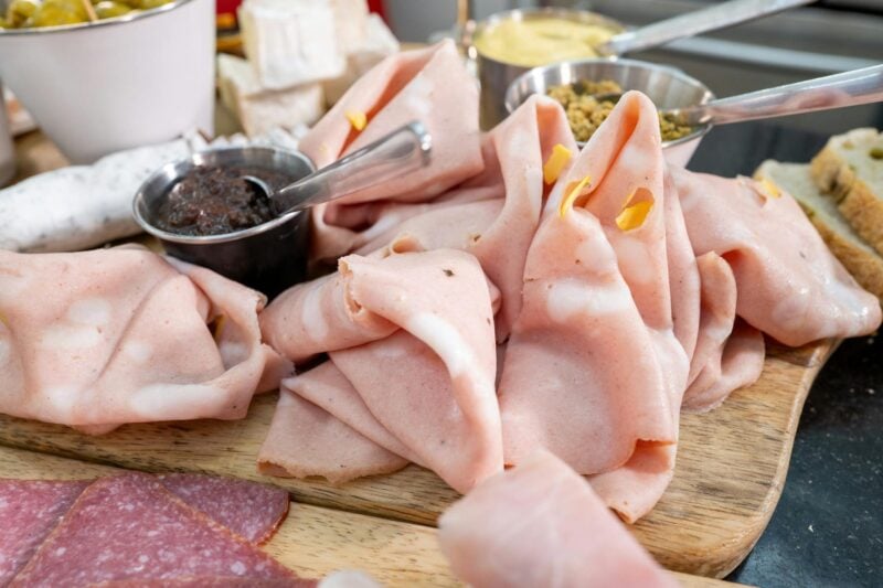 Veganer Aufschnitt in verschiedenen Sorten auf Holzbrett, daneben Brötchen, Senf und Gurke