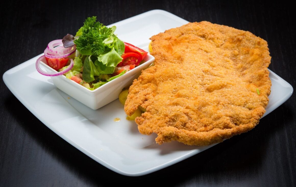 Goldbraun gebackenes veganes Schnitzel auf weißem Teller mit Zitronenscheibe, Petersilie und Kartoffelsalat im Hintergrund