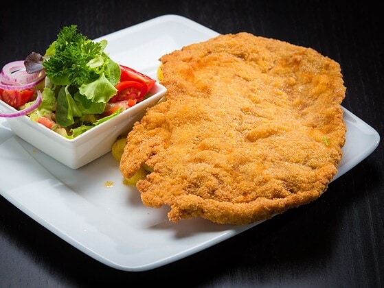 Goldbraun gebackenes veganes Schnitzel auf weißem Teller mit Zitronenscheibe, Petersilie und Kartoffelsalat im Hintergrund