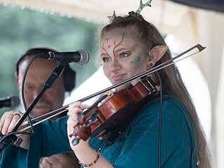 Bardinnen-Auftritt auf der Tavern Stage der Vegan Fantasy Fair: junge Violinistin im Elfen-Cosplay mit Geweih, blau-türkisem Mittelalter-Gewand und kunstvollem Face-Paint spielt Geige, im Hintergrund Begleitgitarrist.