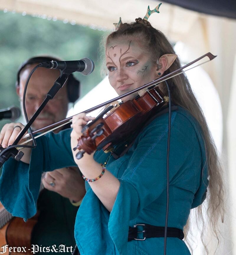 Bardinnen-Auftritt auf der Tavern Stage der Vegan Fantasy Fair: junge Violinistin im Elfen-Cosplay mit Geweih, blau-türkisem Mittelalter-Gewand und kunstvollem Face-Paint spielt Geige, im Hintergrund Begleitgitarrist.