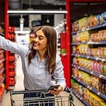 Neue vegane Produkte finden: Mit diesen Tricks bist du immer als Erste dran! Junge Frau mit Smartphone und Einkaufswagen greift im Supermarkt nach einem Produkt