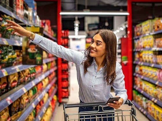Junge Frau mit Smartphone und Einkaufswagen greift im Supermarkt nach einem Produkt