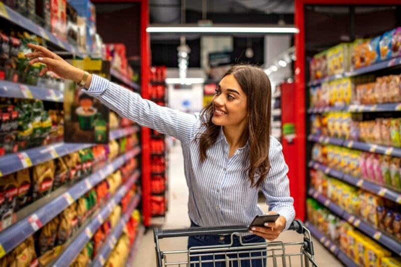 Junge Frau mit Smartphone und Einkaufswagen greift im Supermarkt nach einem Produkt