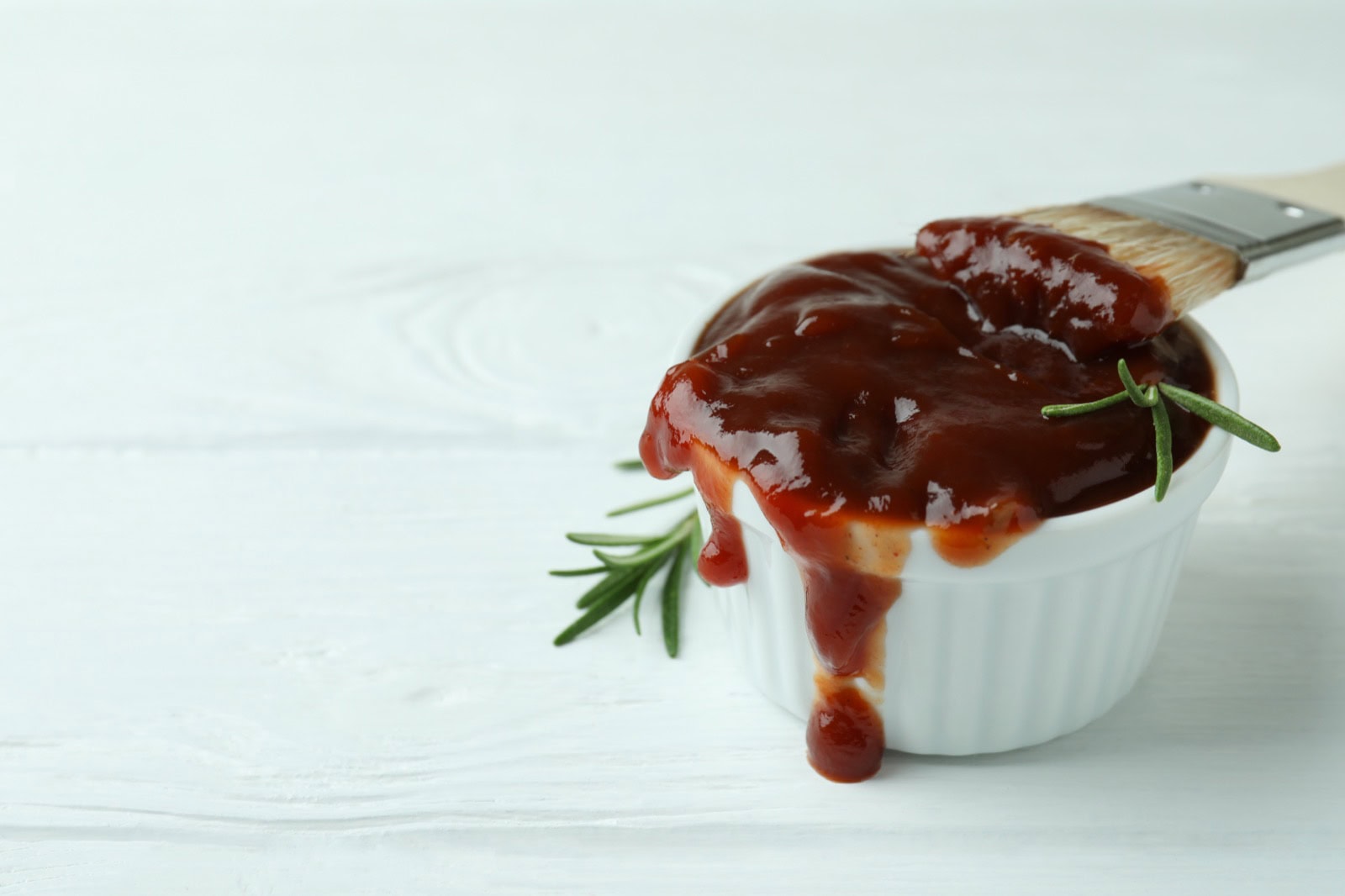 Schüssel mit hausgemachter BBQ-Sauce, Pinsel und Rosmarin auf weißem Holztisch