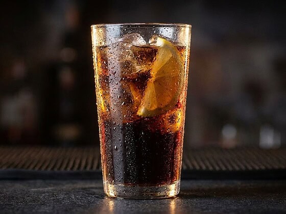 Ist Cola vegan: Glas Cola mit Eiswürfeln und Zitronenscheibe vor dunklem Hintergrund