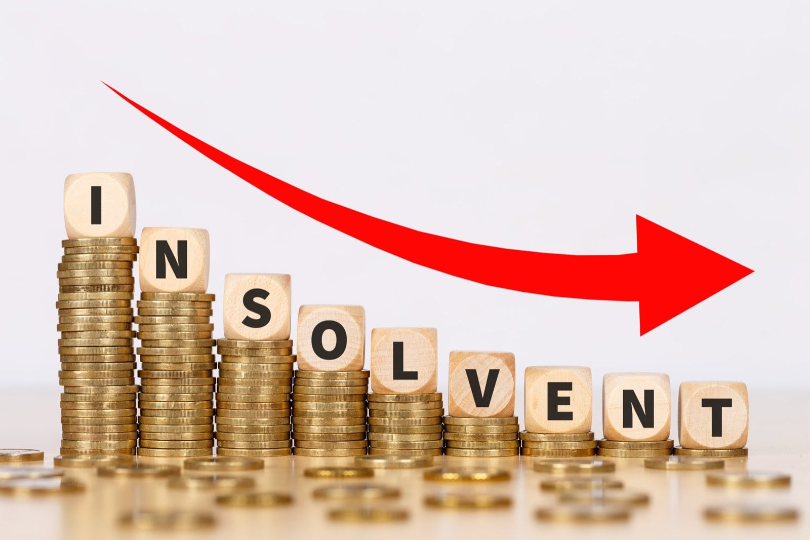 Würfel bilden das Wort Insolvent auf absteigenden Münzstapeln, darüber ein roter Abwärtspfeil